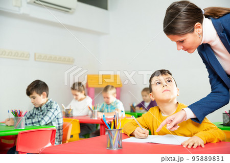 Friendly teacher woman helping boy during lesson in schoolroom Friendly teacher woman helping boy during lesson in schoolroom 95889831