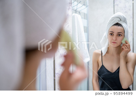 Woman makes a facial massage with a gouache scraper and looks in the mirror. She is wearing a nightie, towel on the head and has bare shoulders Woman makes a facial massage with a gouache scraper and looks in the mirror. She is wearing a nightie, towel on the head and has bare shoulders 95894499