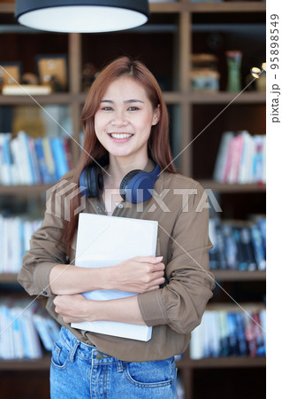A portrait of a young Asian woman with a smiling face looking for a textbook in the library. 95898549