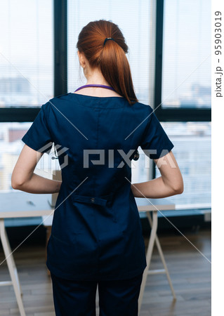 Vertical portrait of unrecognizable female doctor wearing medical uniform standing on background of window. Woman physician with hands at waist posing with stethoscope looking away. 95903019