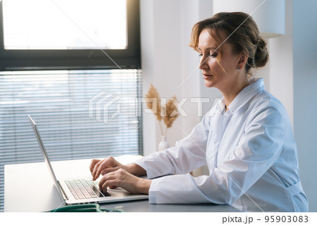 Side view of female doctor physician wearing white coat working typing on laptop computer looking on display screen, sitting at desk in light office room at hospital on background of window. Side view of female doctor physician wearing white coat working typing on laptop computer looking on display screen, sitting at desk in light office room at hospital on background of window. 95903083