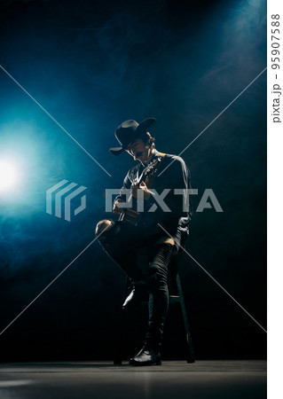 Portrait of man with moustaches in country style clothes playing guitar isolated over dark blue background with smoke. Live performance Portrait of man with moustaches in country style clothes playing guitar isolated over dark blue background with smoke. Live performance 95907588