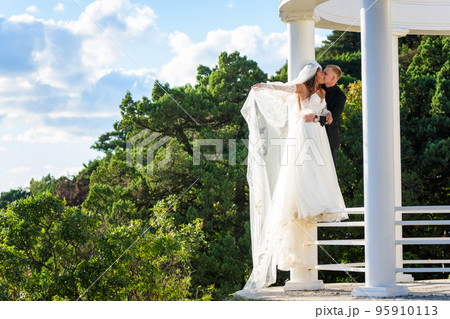The newlyweds happily embrace in a beautiful gazebo with columns against the backdrop of foliage and sky The newlyweds happily embrace in a beautiful gazebo with columns against the backdrop of foliage and sky 95910113