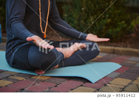 Cropped view of male athlete, yogi wearing rosary beads and gray sportswear, sitting on fitness mat in lotus position and palms up on knees, practicing yoga. Prayer, gratitude. Yoga pose. Meditation. 95911412