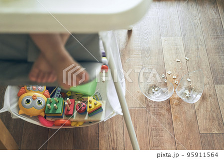 A small baby on a high chair dropped a cup of noodle soup on the wooden floor. The transparent plate is divided into two parts. Noodles in the form of figures. The table is dirty. High quality photo 95911564