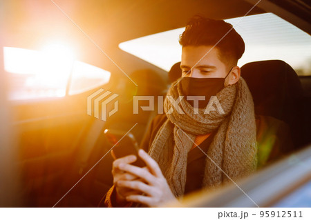 Young man in protective sterile mask in taxi car on a back seat checking her cell phone. 95912511