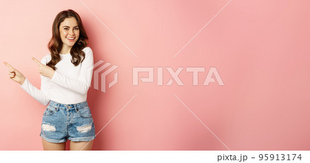 Cute smiling brunette girl, showing advertisement, pointing fingers left at banner, logo on empty pink space, studio background Cute smiling brunette girl, showing advertisement, pointing fingers left at banner, logo on empty pink space, studio background 95913174