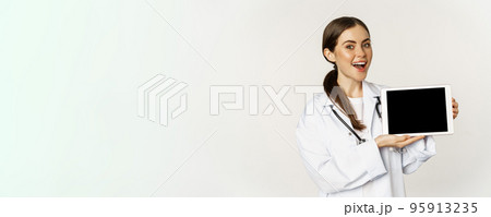 Online medical help, remote appointment. Smiling beautiful woman doctor showing digital tablet screen, demonstrating website logo, standing over white background Online medical help, remote appointment. Smiling beautiful woman doctor showing digital tablet screen, demonstrating website logo, standing over white background 95913235