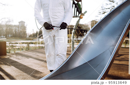 Man wearing protective suit disinfecting the playground in the sun with spray. Covid -19. Cleaning. 95913649