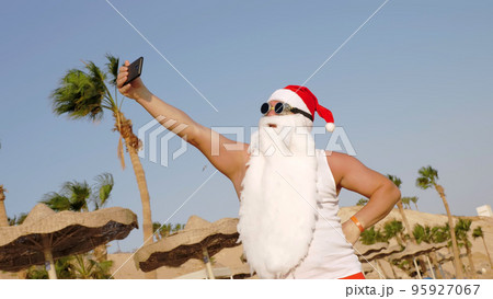 Santa Claus takes selfie. Santa summer vacation. Santa Claus blogger. Funny Santa, in sunglasses, takes selfie video on beach by the sea, against palm trees backdrop. High quality photo 95927067