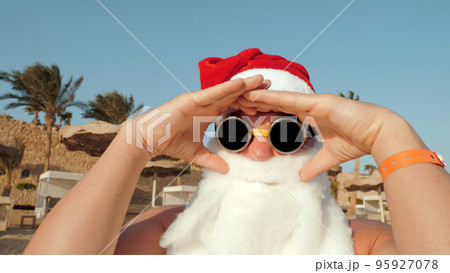 Santa Claus summer vacation. Santa having fun. Funny Santa, in sunglasses, on beach by the sea, against palm trees backdrop. Santa takes selfie. High quality photo 95927078
