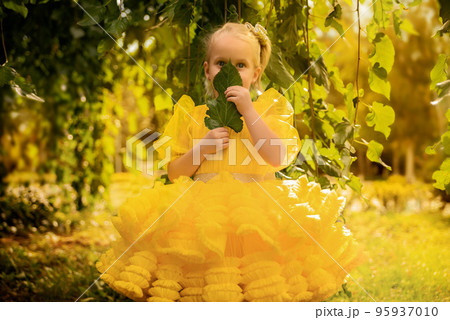 A little girl in a fluffy yellow dress. Little princess with blue eyes among the leaves and bushes 95937010