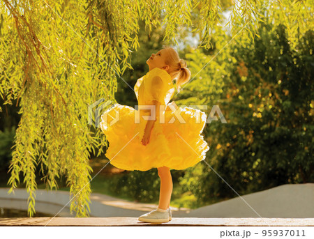 A little girl in a fluffy yellow dress. Little princess with blue eyes among the leaves and bushes 95937011
