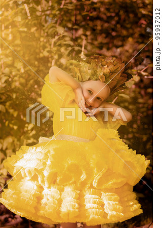 A little girl in a fluffy yellow dress. Little princess with blue eyes among the leaves and bushes 95937012