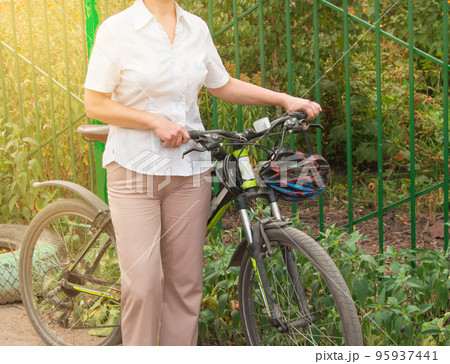 Middle-aged attractive slim woman in light trousers and shirt stands near the bike in the Park on a Sunny summer day, Cycling concept for adult women Middle-aged attractive slim woman in light trousers and shirt stands near the bike in the Park on a Sunny summer day, Cycling concept for adult women 95937441