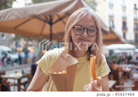 Close-up of young blonde woman eating eating traditional spanish snack named Churros with sugar and chocolate sauce, a fried-dough pastry based snack. Close-up of young blonde woman eating eating traditional spanish snack named Churros with sugar and chocolate sauce, a fried-dough pastry based snack. 95937469