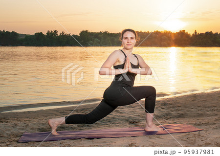 A woman practices yoga while standing in the position of Warrior 1 or Virabhadrasana One. Yoga at sunset in nature near the river. A woman practices yoga while standing in the position of Warrior 1 or Virabhadrasana One. Yoga at sunset in nature near the river. 95937981
