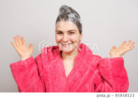 A woman applies a mask to her hair and wraps a film around her head, smiling in a pink bathrobe. The concept of hair care. 95938523