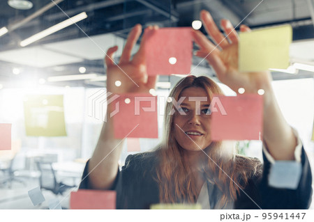 Close up of businesswoman is glues sticky notes on board in modern office Close up of businesswoman is glues sticky notes on board in modern office 95941447