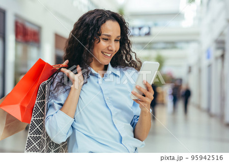 Happy and successful woman shopping for clothes in a supermarket store, Hispanic woman holding a smartphone reading online messages and browsing offers with discounts and sales Happy and successful woman shopping for clothes in a supermarket store, Hispanic woman holding a smartphone reading online messages and browsing offers with discounts and sales 95942516