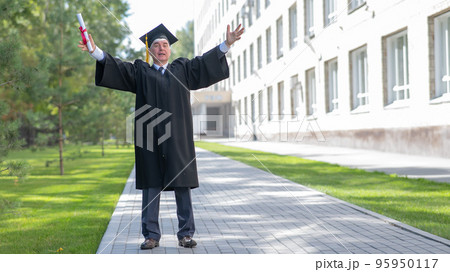 An elderly gray-haired man in a graduate robe spread his arms to the sides and holds a diploma outdoors. An elderly gray-haired man in a graduate robe spread his arms to the sides and holds a diploma outdoors. 95950117