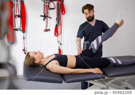 Chiropractor stretches a female patient leg. Male physiotherapist is helping woman stretching his leg in exercise room Chiropractor stretches a female patient leg. Male physiotherapist is helping woman stretching his leg in exercise room 95962209