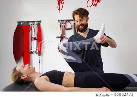 Side view of a cheerful physiotherapist doctor and his female patient who has problems with her leg. Rehabilitation specialist bending woman's knees Side view of a cheerful physiotherapist doctor and his female patient who has problems with her leg. Rehabilitation specialist bending woman's knees 95962472