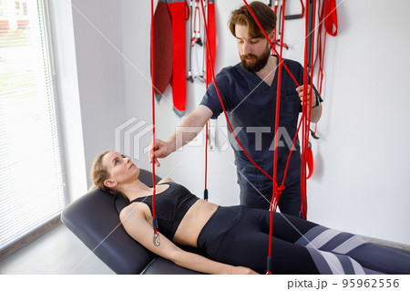 Physiotherapist doctor assists female patient with injury undergoing rehabilitative physiotherapy on a suspension rope system. Modern clinic using advanced techniques rehabilitating procedures Physiotherapist doctor assists female patient with injury undergoing rehabilitative physiotherapy on a suspension rope system. Modern clinic using advanced techniques rehabilitating procedures 95962556