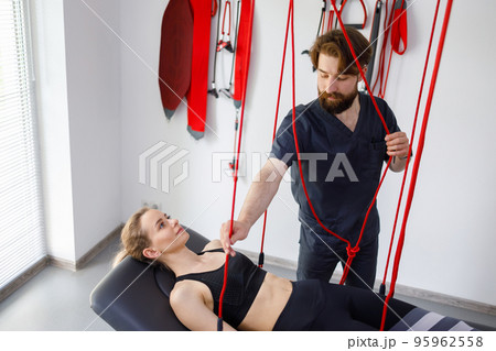 Physiotherapist assists female patient undergoing rehabilitative physiotherapy on a suspension rope system. Modern clinic using advanced rehabilitating therapy 95962558