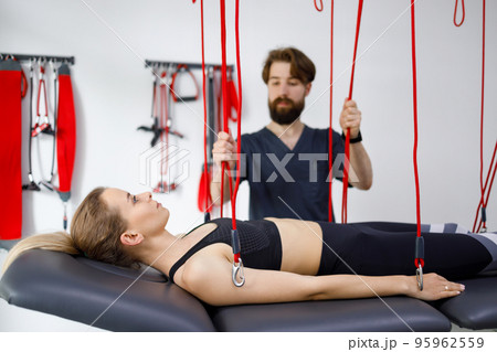 Physiotherapist assists female patient undergoing rehabilitative physiotherapy on a suspension rope system. Modern clinic using advanced rehabilitating therapy Physiotherapist assists female patient undergoing rehabilitative physiotherapy on a suspension rope system. Modern clinic using advanced rehabilitating therapy 95962559