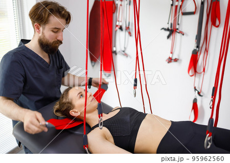 Physiotherapist assists female patient undergoing rehabilitative physiotherapy on a suspension rope system. Modern clinic using advanced rehabilitating therapy Physiotherapist assists female patient undergoing rehabilitative physiotherapy on a suspension rope system. Modern clinic using advanced rehabilitating therapy 95962560