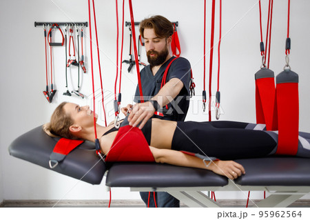 Physiotherapist assists female patient undergoing rehabilitative physiotherapy on a suspension rope system. Modern clinic using advanced rehabilitating therapy 95962564