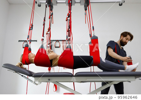 Physiotherapist assists female patient undergoing rehabilitative physiotherapy on a suspension rope system. Modern clinic using advanced rehabilitating therapy Physiotherapist assists female patient undergoing rehabilitative physiotherapy on a suspension rope system. Modern clinic using advanced rehabilitating therapy 95962646