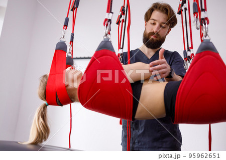Physiotherapist assists female patient undergoing rehabilitative physiotherapy on a suspension rope system. Modern clinic using advanced rehabilitating therapy 95962651