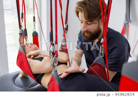 Physiotherapist assists female patient undergoing rehabilitative physiotherapy on a suspension rope system. Modern clinic using advanced rehabilitating therapy 95962654