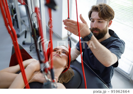 Physiotherapist doctor kneads the neck of a young female patient hanging on the slings of a suspension system for rehabilitation and neuromuscular activation 95962664