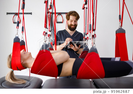 Physiotherapist doctor with clipboard watching female patient hanging on suspension system for neuromuscular activation in rehabilitation clinic Physiotherapist doctor with clipboard watching female patient hanging on suspension system for neuromuscular activation in rehabilitation clinic 95962687