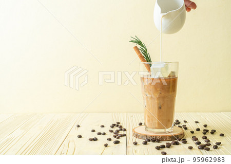pouring milk in black coffee glass with ice cube, cinnamon and rosemary 95962983