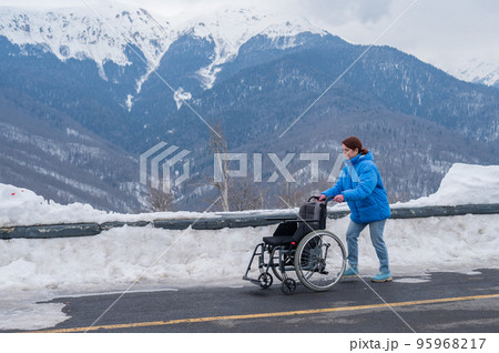 Caucasian woman carries an empty wheelchair among the snowy mountains.  95968217