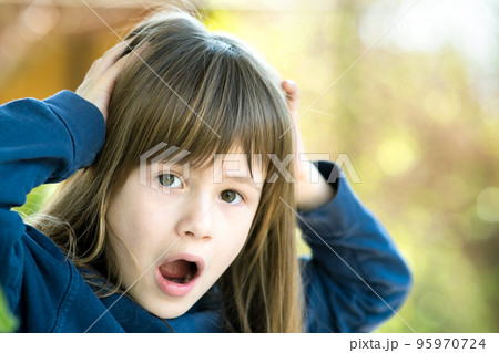Portrait of surprised child girl holding hands to her head outdoors in summer. Shocked female kid on a warm day outside. Portrait of surprised child girl holding hands to her head outdoors in summer. Shocked female kid on a warm day outside. 95970724