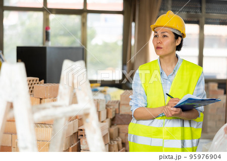 Female foreman in a protective helmet and vest checks the completed work on the repair of room Female foreman in a protective helmet and vest checks the completed work on the repair of room 95976904