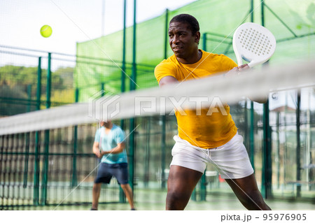 Male players playing padel in a padel court outdoor behind net Male players playing padel in a padel court outdoor behind net 95976905