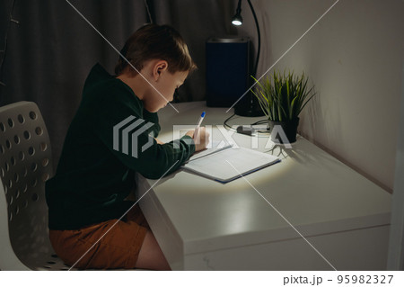 caucasian boy doing homework at home in dark room by the light of lamp. 95982327