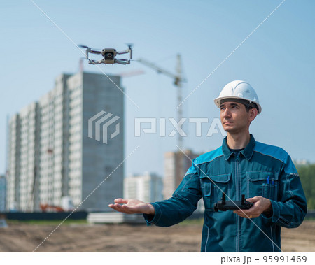 A man in a helmet and overalls controls a drone at a construction site. The builder carries out technical oversight. A man in a helmet and overalls controls a drone at a construction site. The builder carries out technical oversight. 95991469