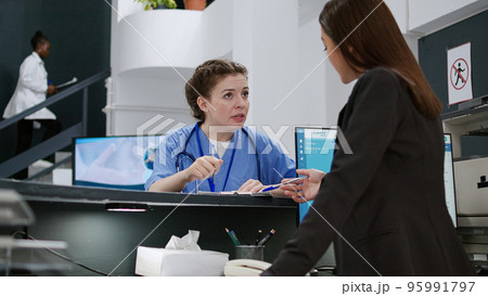 Portrait of medical assistant talking to receptionist at hospital registration counter, using appointment reports to help patients with checkup visits. Working at reception desk. Handheld shot. Portrait of medical assistant talking to receptionist at hospital registration counter, using appointment reports to help patients with checkup visits. Working at reception desk. Handheld shot. 95991797
