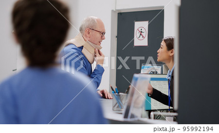 Old man with medical neck collar asking for help at hospital reception desk, talking to asian receptionist and medical assistant. Nurse taking patient with cervical foam at checkup visit, injury pain. Old man with medical neck collar asking for help at hospital reception desk, talking to asian receptionist and medical assistant. Nurse taking patient with cervical foam at checkup visit, injury pain. 95992194
