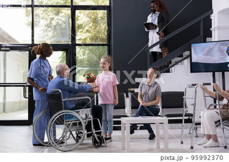 Granddaughter handing bouquet of flowers to disabled grandfather after finishing medical consultation in hospital waiting area. Elderly man discussing health care treatment with nurse 95992370