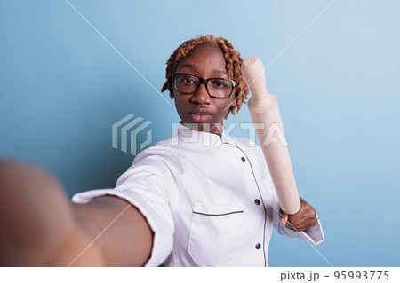 Angry female dinning hall subordinate threatening via video call. Annoyed african american chef intimidating with rolling pin. Self portrait of angry cooker in uniform. Angry female dinning hall subordinate threatening via video call. Annoyed african american chef intimidating with rolling pin. Self portrait of angry cooker in uniform. 95993775