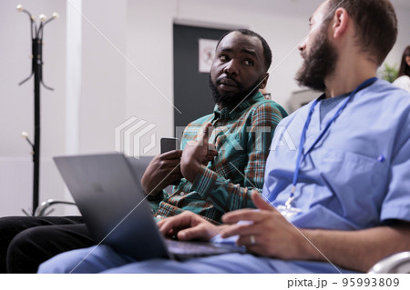 African american concerned patient talking to nurse. Hospital medical staff member discussing young man health status. Different nationalities people talking seriously in medical tower reception area. African american concerned patient talking to nurse. Hospital medical staff member discussing young man health status. Different nationalities people talking seriously in medical tower reception area. 95993809