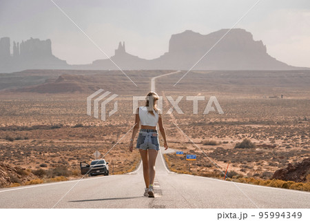 Rear view of woman walking on road leading towards Monument valley. Female tourist is spending leisure time in desert with car parked at roadside. She is exploring at Navajo Tribal Park during summer. 95994349
